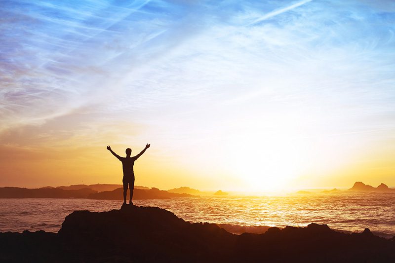 A person in addiction recovery raises their arms in happiness while watching the sunset.